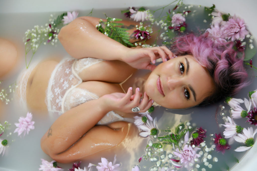 woman with pink hair wearing a white lace bra and panty set posing in a milk bath with purple, white, and green flowers