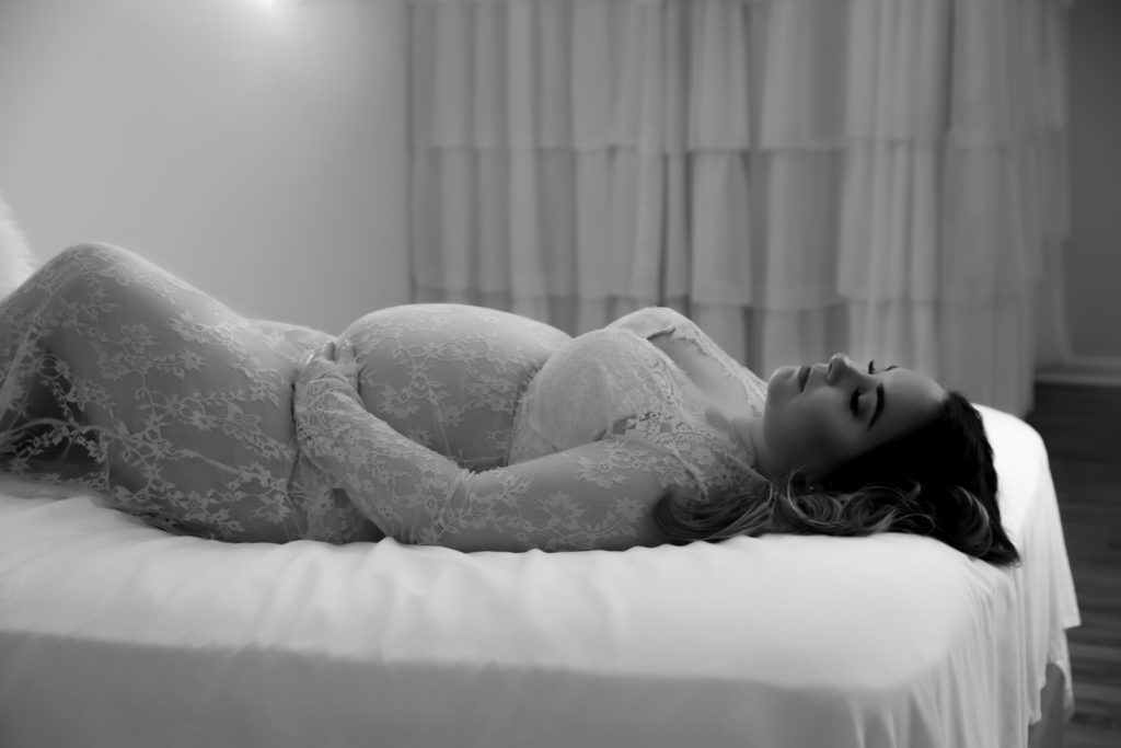 pregnant woman wearing a nude full length lace bodysuit posing on a bed